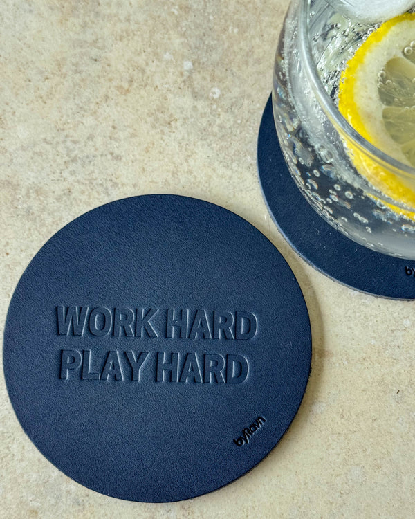 MOKKA glassbrikke i mørkeblått skinn preget "Work hard play hard" på lyst bord ved glass med sitronskive
