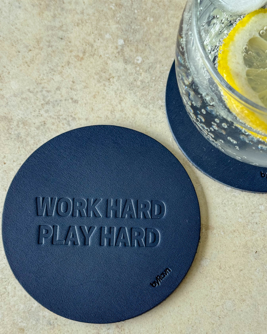 MOKKA glassbrikke i mørkeblått skinn preget "Work hard play hard" på lyst bord ved glass med sitronskive
