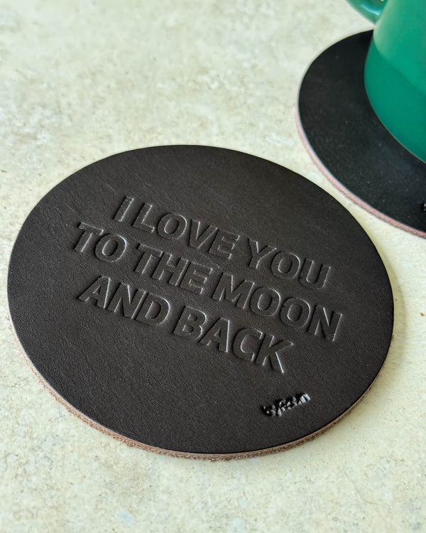 MOKKA glassbrikke i mørkebrunt skinn preget "I love you to the moon and back" på lyst bord ved grønn kopp