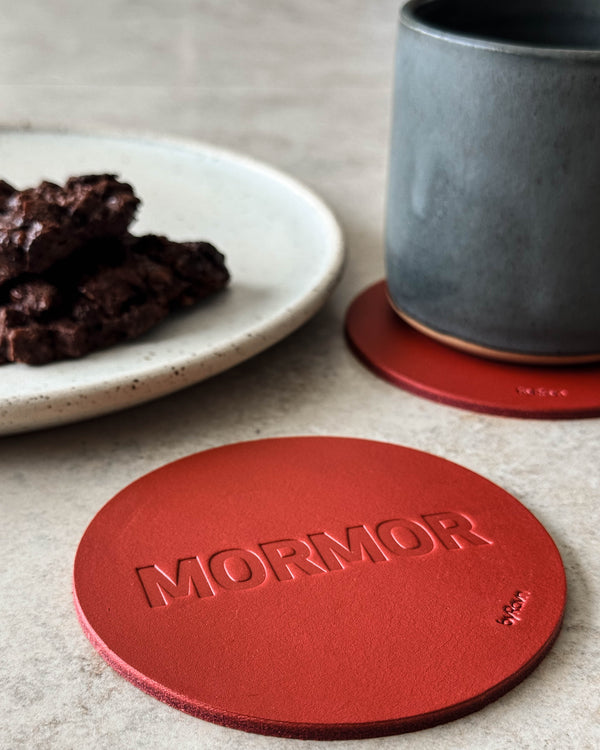 MOKKA glassbrikke i rødt skinn preget "Mormor" på lyst bord med grå kaffekopp og tallerken med sjokoladekake