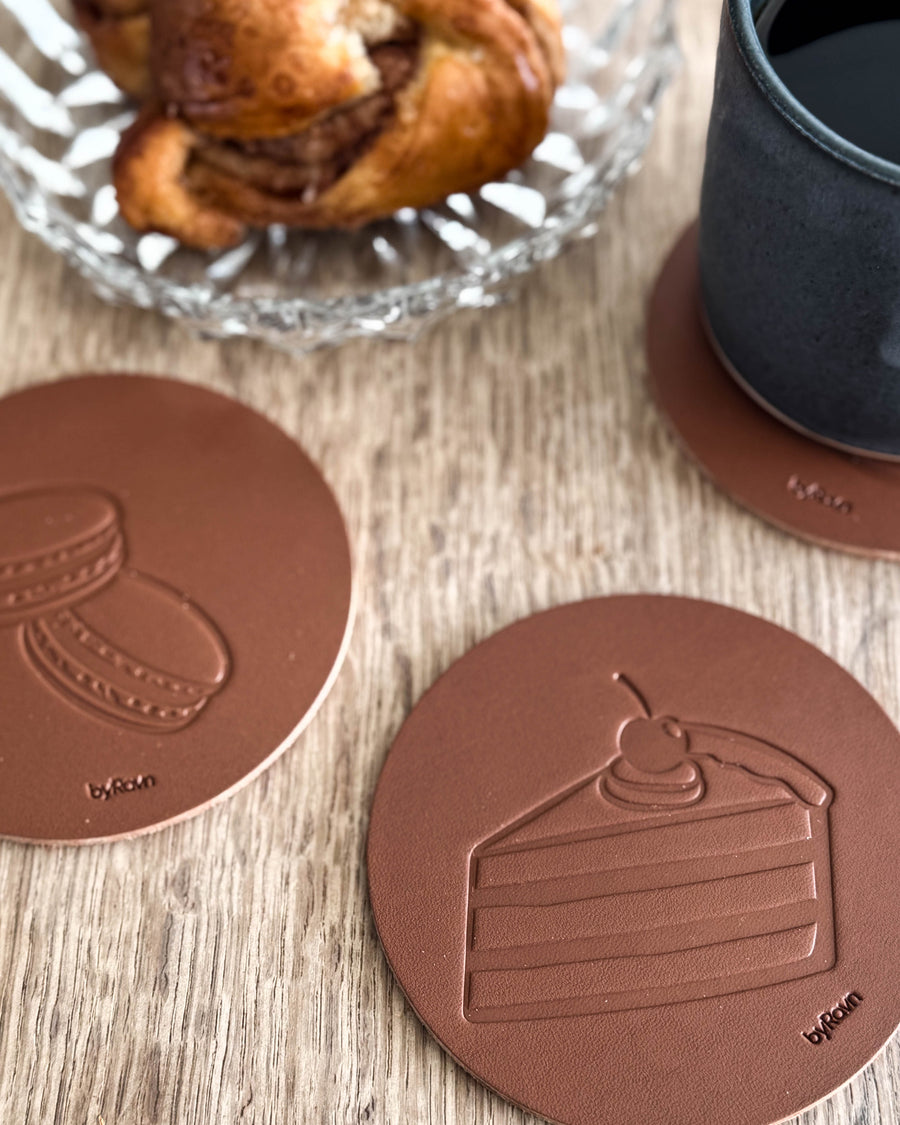 MOKKA kaffe & kake-glassbrikker i cognac skinn preget med bløtkake og macarons på trebord ved kanelsnurr og svart kopp