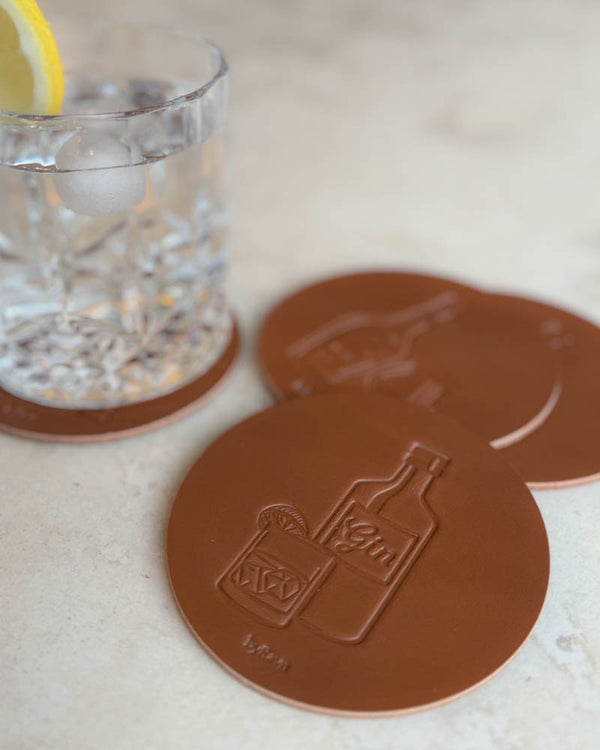 MOKKA brennevin-glassbrikker i cognac skinn preget med ginflaske og drink, på lys steinflate ved glass med sitronskive