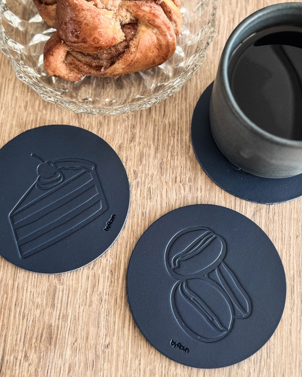 MOKKA kaffe & kake-glassbrikker i mørkeblått skinn preget med bløtkake og kaffebønner på trebord ved kanelsnurr og grå kopp
