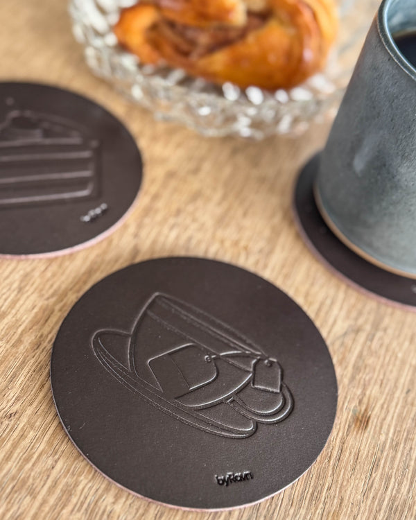 MOKKA kaffe & kake-glassbrikker i mørkebrunt skinn preget med tekopp og bløtkake på trebord ved kanelsnurr og grønn kopp
