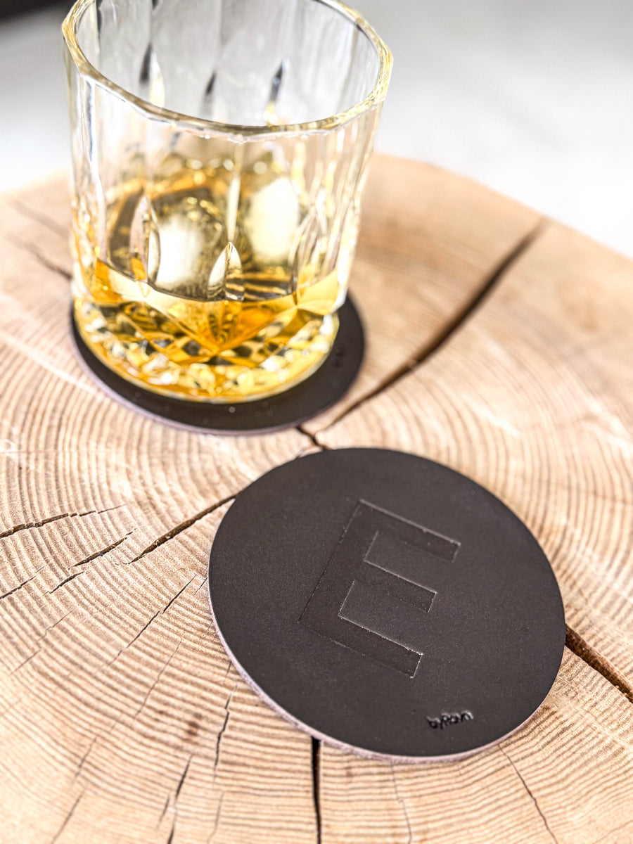 MOKKA glassbrikke i mørkebrunt skinn preget med bokstaven "E" på treskive ved whiskyglass på glassbrikke
