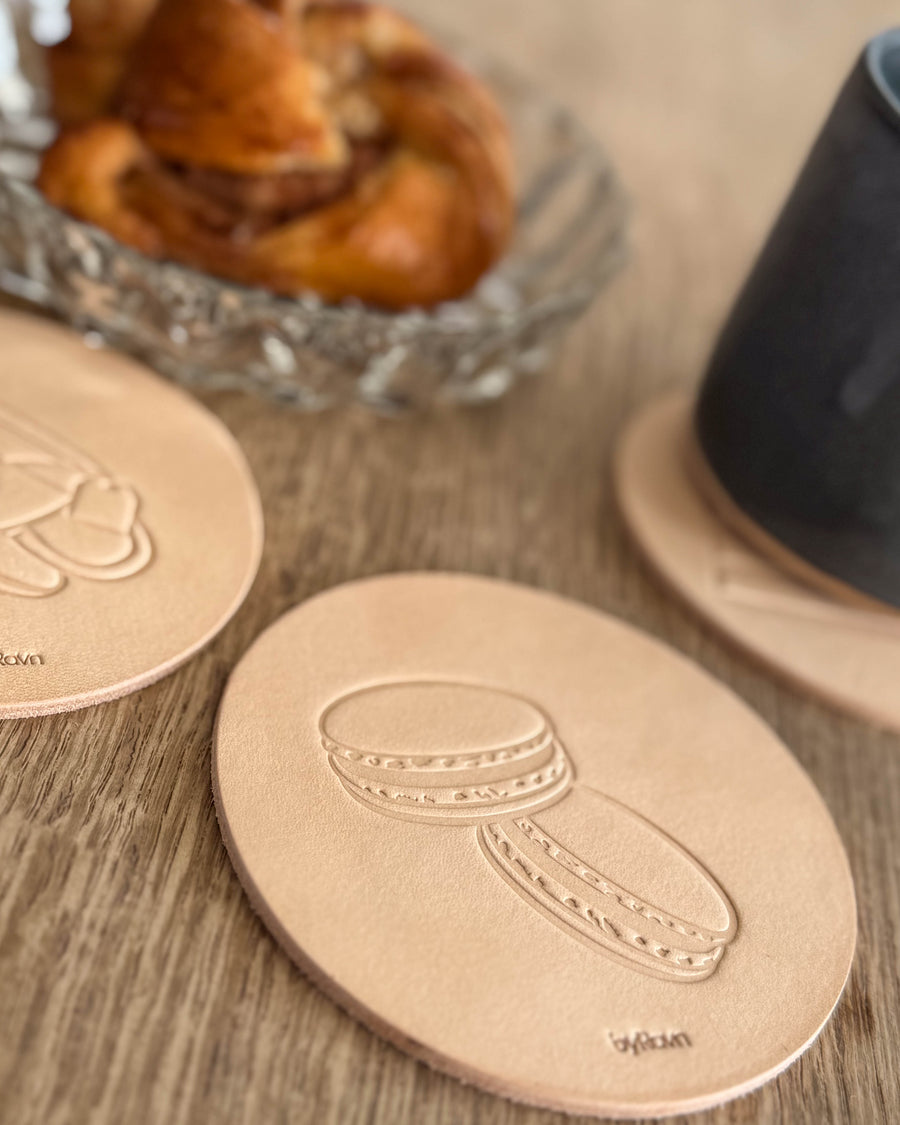 MOKKA kaffe & kake-glassbrikke i naturfarget kjerneskinn preget med macarons på trebord ved kanelsnurr og svart kopp