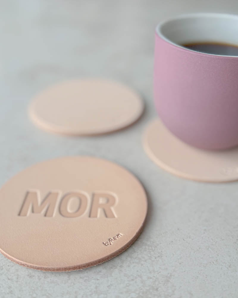 MOKKA glassbrikke i naturfarget kjerneskinn preget "Mor" i store bokstaver, rosa kaffekopp på glassbrikke bak