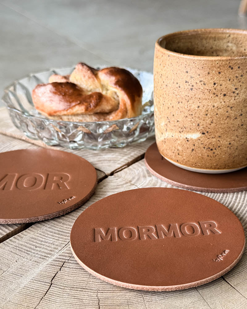 MOKKA glassbrikker i cognac skinn preget "Mor" og "Mormor" på trebord med kaffekopp og kanelsnurr i glasskål
