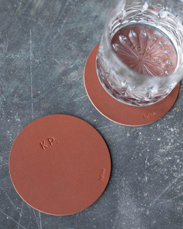 MOKKA runde glassbrikker i cognac skinn med initialene "K.P." gravert, én under krystallglass på grått betongbord