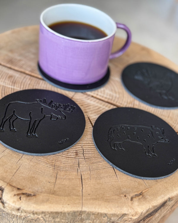 MOKKA jakt-glassbrikker i svart skinn preget med elg, villsvin og tiur på treskive med lilla kaffekopp på glassbrikke