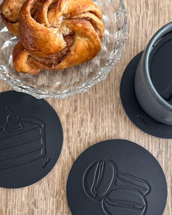 MOKKA kaffe & kake-glassbrikker i svart skinn preget med bløtkake og kaffebønner på trebord ved kanelsnurr og grå kopp