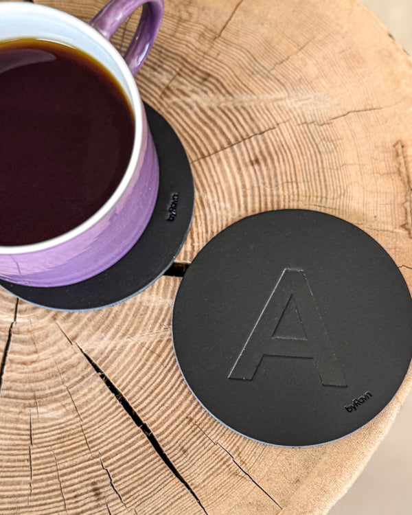 MOKKA glassbrikke i svart skinn preget med bokstaven "A" på treskive ved lilla kaffekopp på glassbrikke