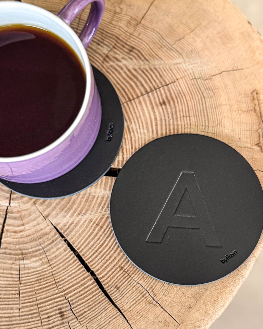 MOKKA glassbrikke i svart skinn preget med bokstaven "A" på treskive ved lilla kaffekopp på glassbrikke