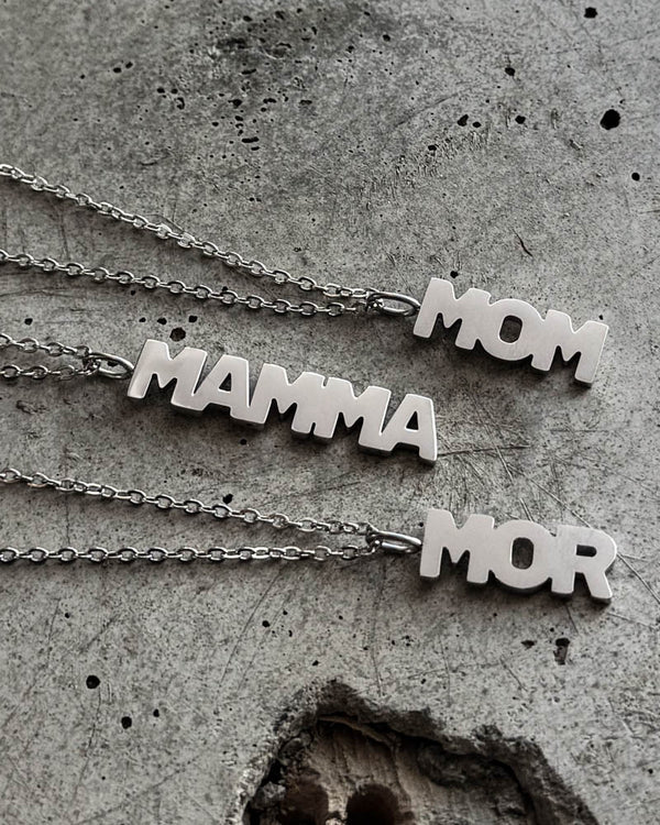 MOM halskjeder i sølvfarget rustfritt stål med bokstavanheng "MOM", "MAMMA" og "MOR", liggende på rå betong