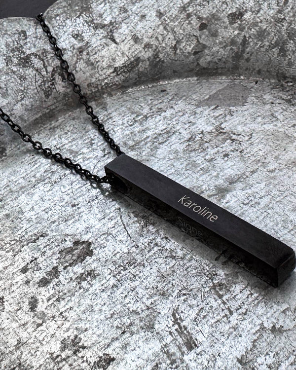 NEO navnehalskjede i svart stål med navnet "Karoline" gravert på avlang stav, liggende på patinert sinkfat