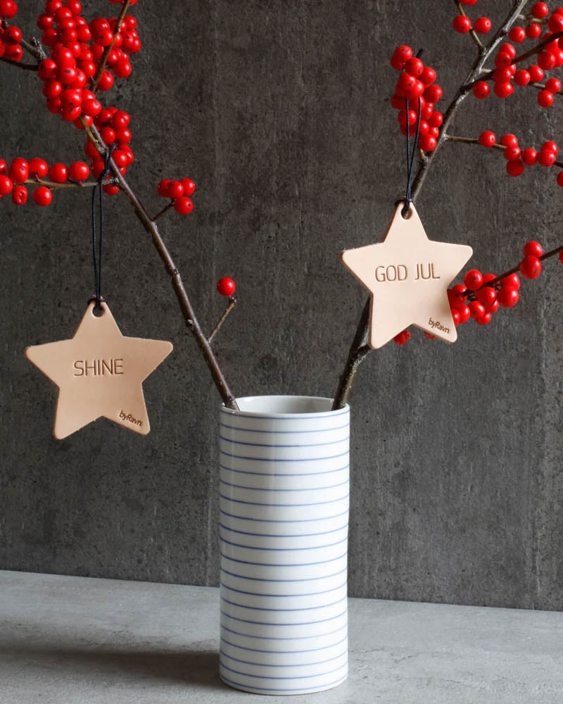 NORTH julestjerner i naturfarget skinn med graveringene "Shine" og "God jul" hengt på greiner med røde bær i stripet vase
