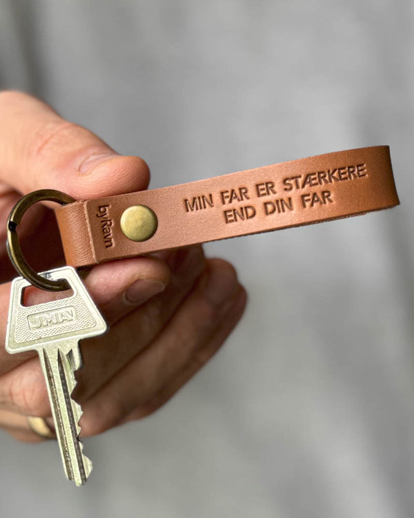 RAVN nøkkelring i cognac skinn med graveringen "Min far er stærkere end din far", holdt i hånden med nøkkel