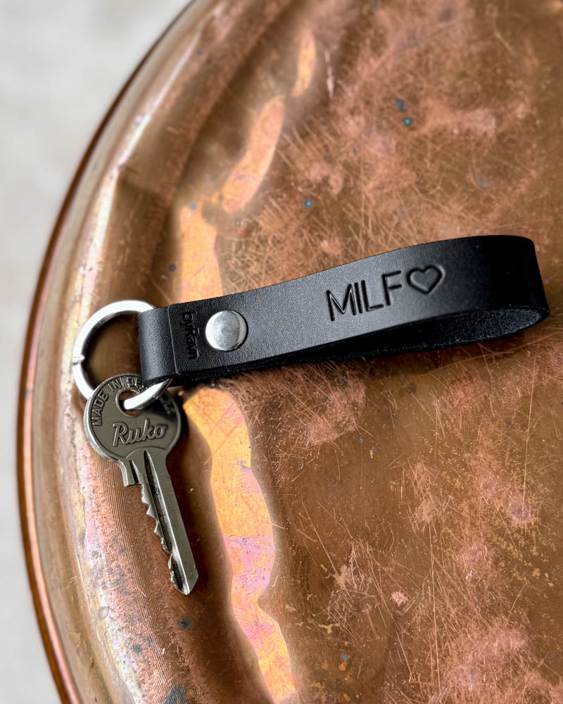 RAVN nøkkelring i mørkt skinn med graveringen "MILF" ♡ og nøkkel, liggende på patinert kobberbrett