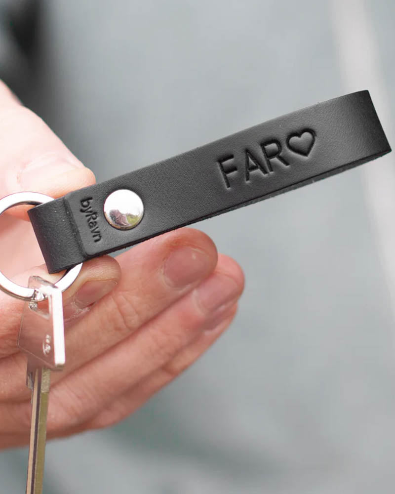 RAVN nøkkelring i svart skinn med graveringen "Far" ♡ og nagle i stål, holdt i hånden med nøkkel