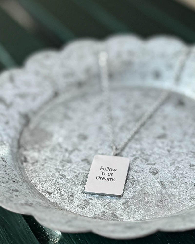 RAZ halskjede i sølvfarget rustfritt stål med graveringen "Follow Your Dreams" på rektangulær plate, på hvitpatinert sinkfat