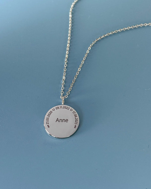 SIA CIRCLE navnehalskjede i sølvfarget rustfritt stål med "Anne" og datoene "21.03.2020 ♡ 09.11.2022 ♡ 17.06.2025" i sirkel, på blå bunn