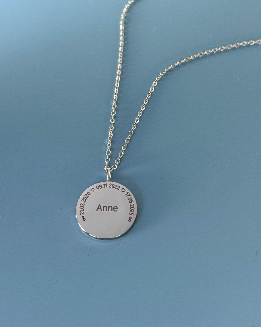 SIA CIRCLE navnehalskjede i sølvfarget rustfritt stål med "Anne" og datoene "21.03.2020 ♡ 09.11.2022 ♡ 17.06.2025" i sirkel, på blå bunn