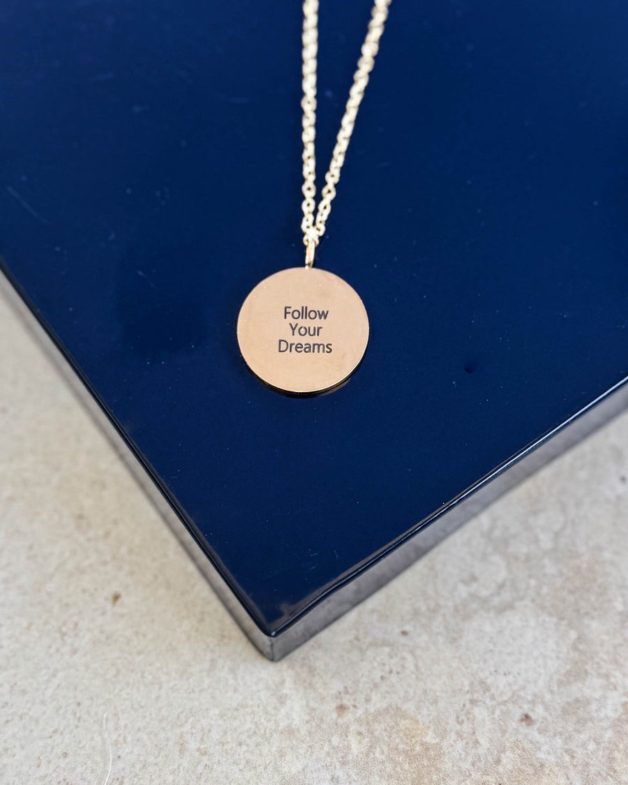 SIA QUOTE halskjede i gullbelagt stål med graveringen "Follow your dreams" på rundt anheng, på mørkeblå eske