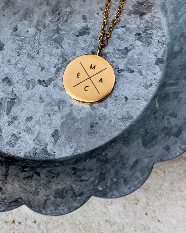SIA X halskjede i gullbelagt stål med initialene "M E A C" gravert i kryss på rundt anheng, liggende på sinkfat