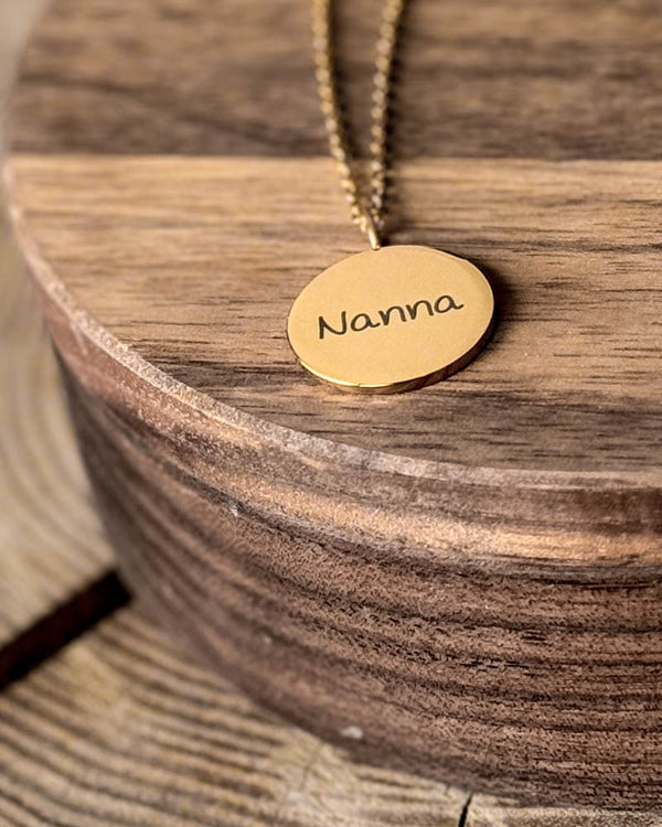 SIA NOTE SCRIPT navnehalskjede i gullbelagt stål med navnet "Nanna" i håndskrift gravert på rundt anheng, på rundt trefat
