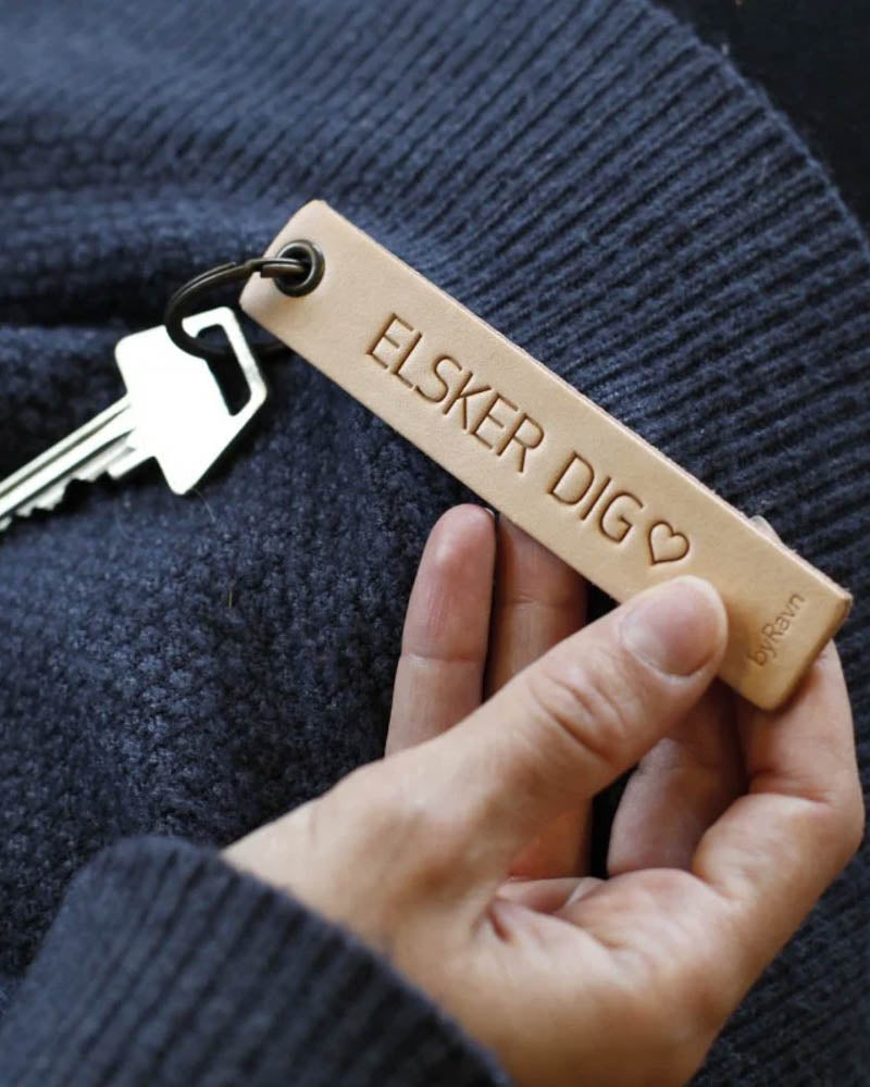 SOLO Medium nøkkelring i naturfarget skinn holdt i hånden med nøkkel og graveringen "Elsker dig" ♡