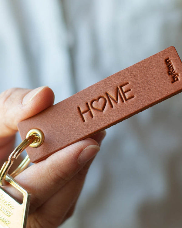 SOLO Small nøkkelring i cognac skinn holdt i hånden med nøkkel og graveringen "Home", der O-en er et hjerte