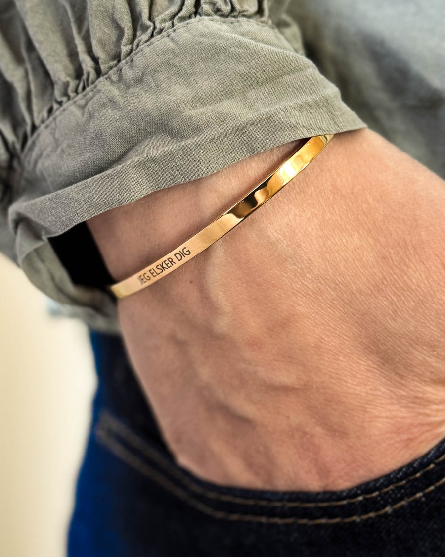 VENUS armbånd i gullbelagt stål med graveringen "Jeg elsker dig" rundt håndleddet, hånd i lommen på mørke jeans