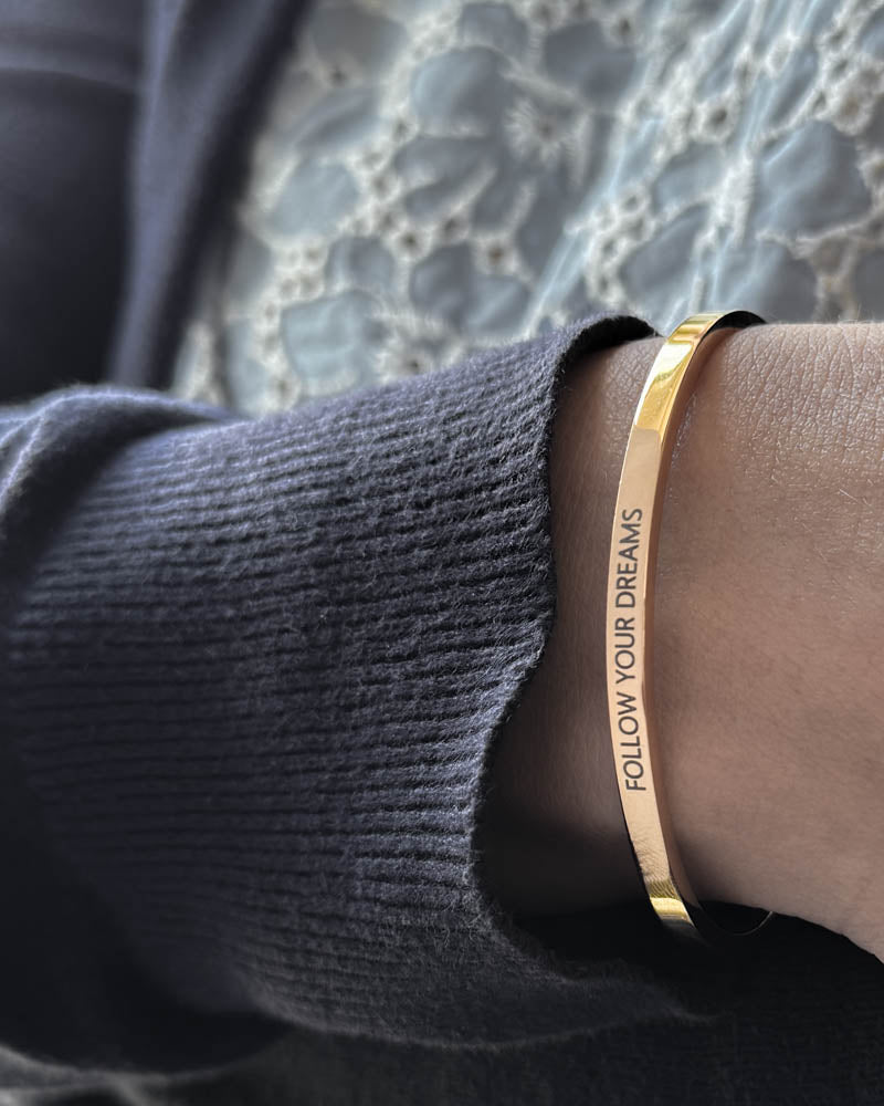 VENUS armbånd i gullbelagt stål med graveringen "Follow your dreams" i versaler, båret til mørkeblå strikk og blondebluse
