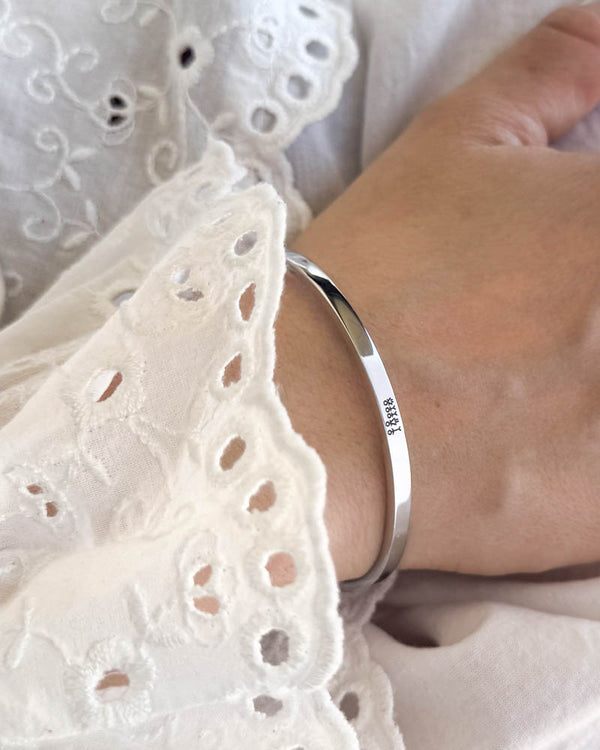 VENUS STICK armbånd i sølvfarget rustfritt stål med gravert fyrstikkfamilie, båret til hvit bluse med broderi